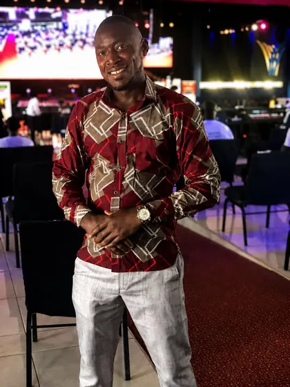 Valentine Golden Ghanem - Portrait at an event venue, patterned maroon shirt, stage screens behind him.