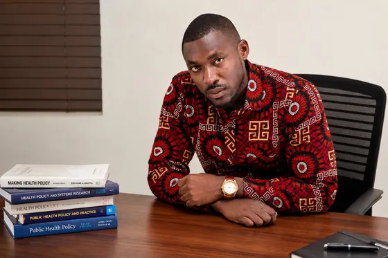 Valentine Golden Ghanem - At his desk with health-policy textbooks, red patterned shirt.