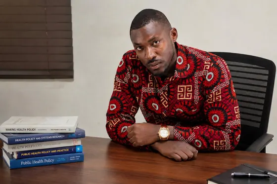 Valentine Golden Ghanem - At his desk, arms resting beside health-policy texts, red patterned shirt.