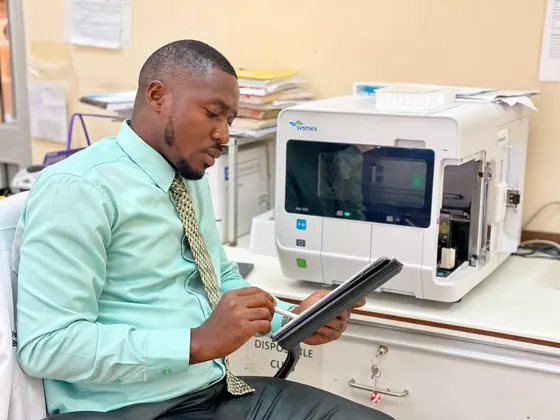 Valentine Golden Ghanem - Beside a laboratory analyser, mint-green shirt, reviewing a tablet.