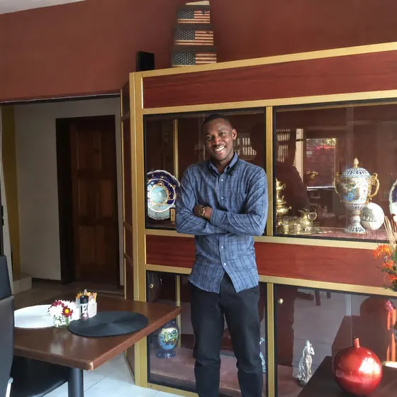 Valentine Golden Ghanem - Portrait at home, beside a display cabinet, navy checkered shirt.