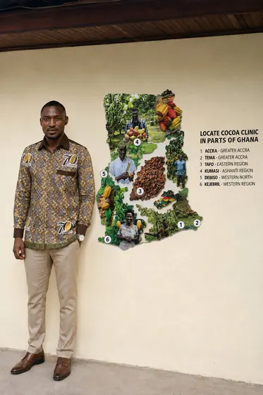 Valentine Golden Ghanem - Beside a wall-mounted map of Cocoa Clinic locations, Ghana COCOBOD 70th Anniversary shirt.