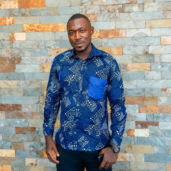 Valentine Golden Ghanem - Portrait against a stone wall, navy patterned shirt, phone in hand.